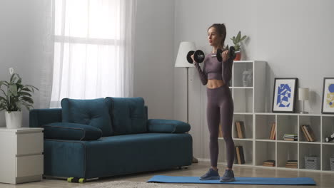 young-sporty-woman-is-training-with-dumbbells-standing-in-living-room-at-daytime-full-length-portrait-of-sportswoman-in-modern-interior